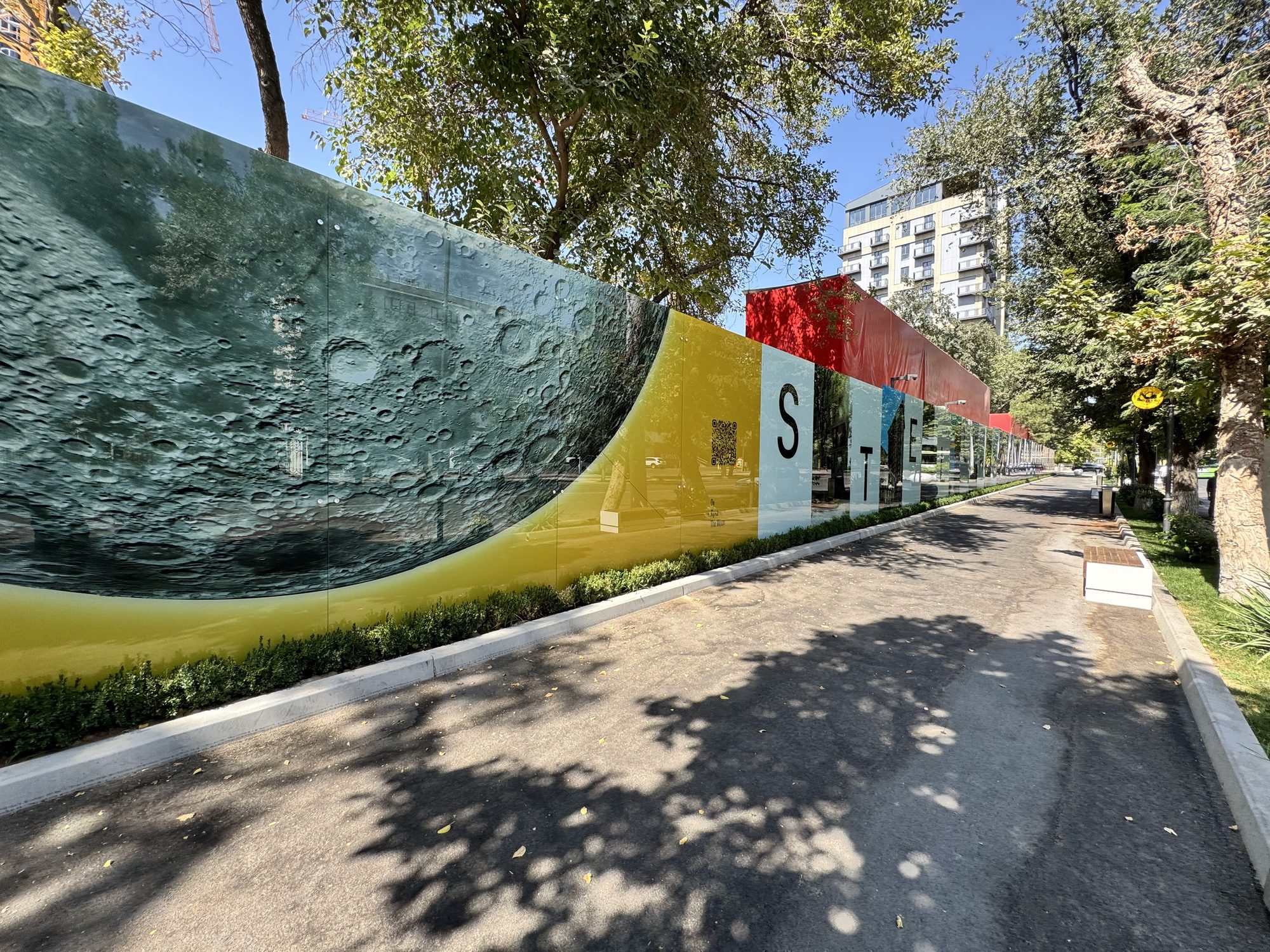 WE WALL printed glass construction hoarding — full panel run with space-themed artwork along a tree-lined street in Tashkent